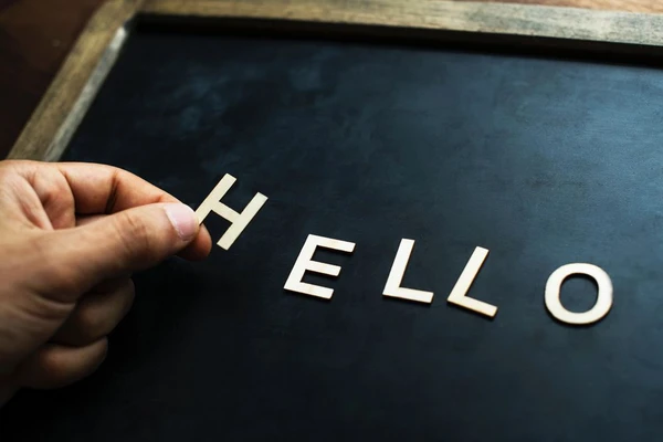 Egy kéz fehér H betűt helyez a HELLO szó elejére egy sötét táblán, amelyet világos fakeret vesz körül.